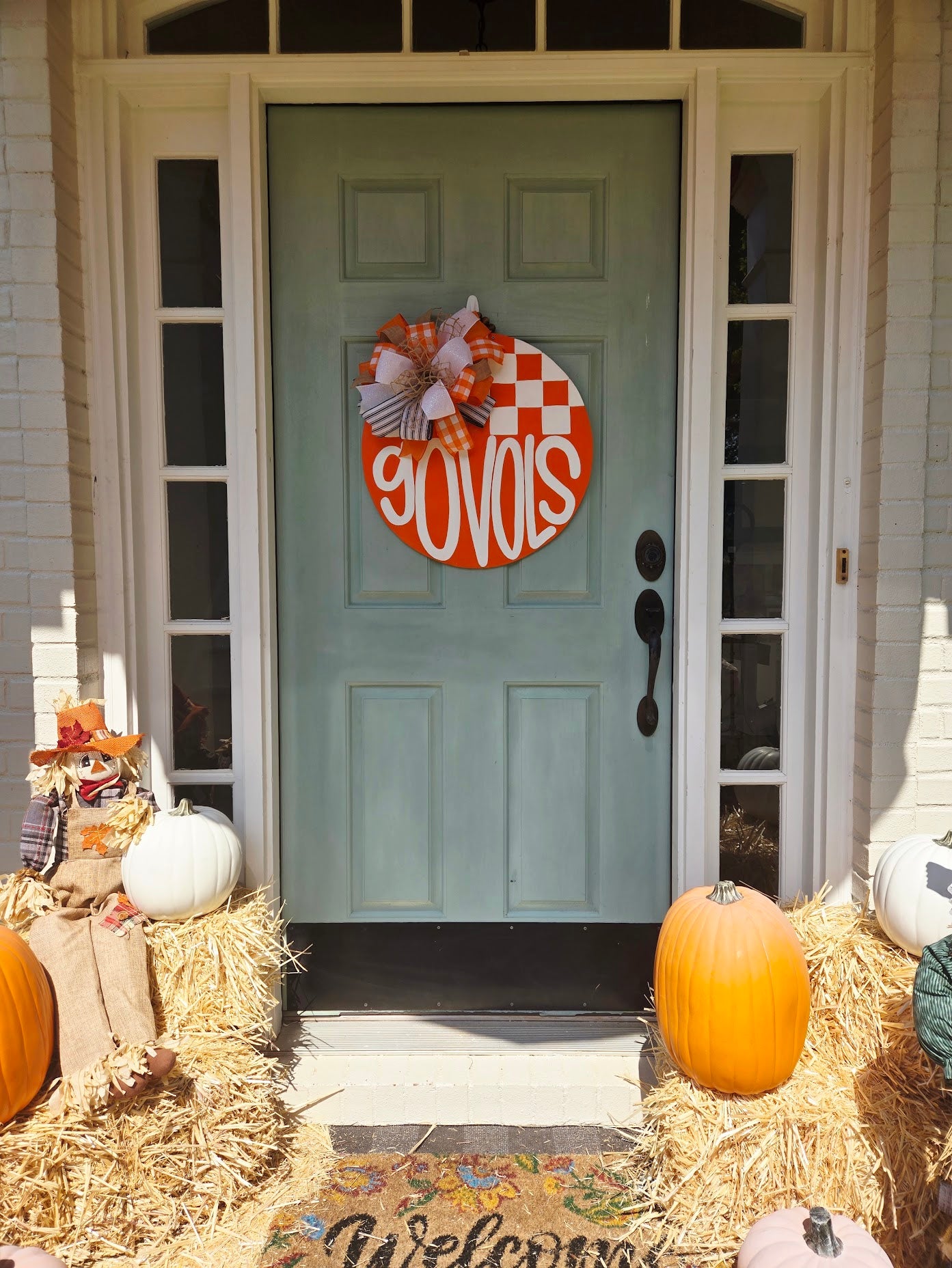 A round orange and white checkered fall wreath with the word 'COUSIS' in white script, hung on a light blue front door. The porch is decorated with pumpkins, hay bales, and a straw scarecrow.